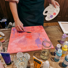Person verteilt Acrylfarben mit Spachtel auf pink grundierter Leinwand beim Abstract Painting Workshop von GRIMMS GARTEN Berlin
