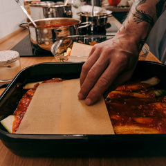 Hand schichtet Lasagneplatten auf Soße, Kochutensilien im Hintergrund beim Batch Cooking Workshop von GRIMMS GARTEN, Meal Prep Kochkurs in Berlin