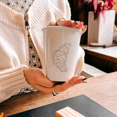 Zwei Hände halten Keramik To-Go Becher mit Croissant-Skizze beim Keramik bemalen Workshop von GRIMMS GARTEN, DIY Kreativkurs in Berlin