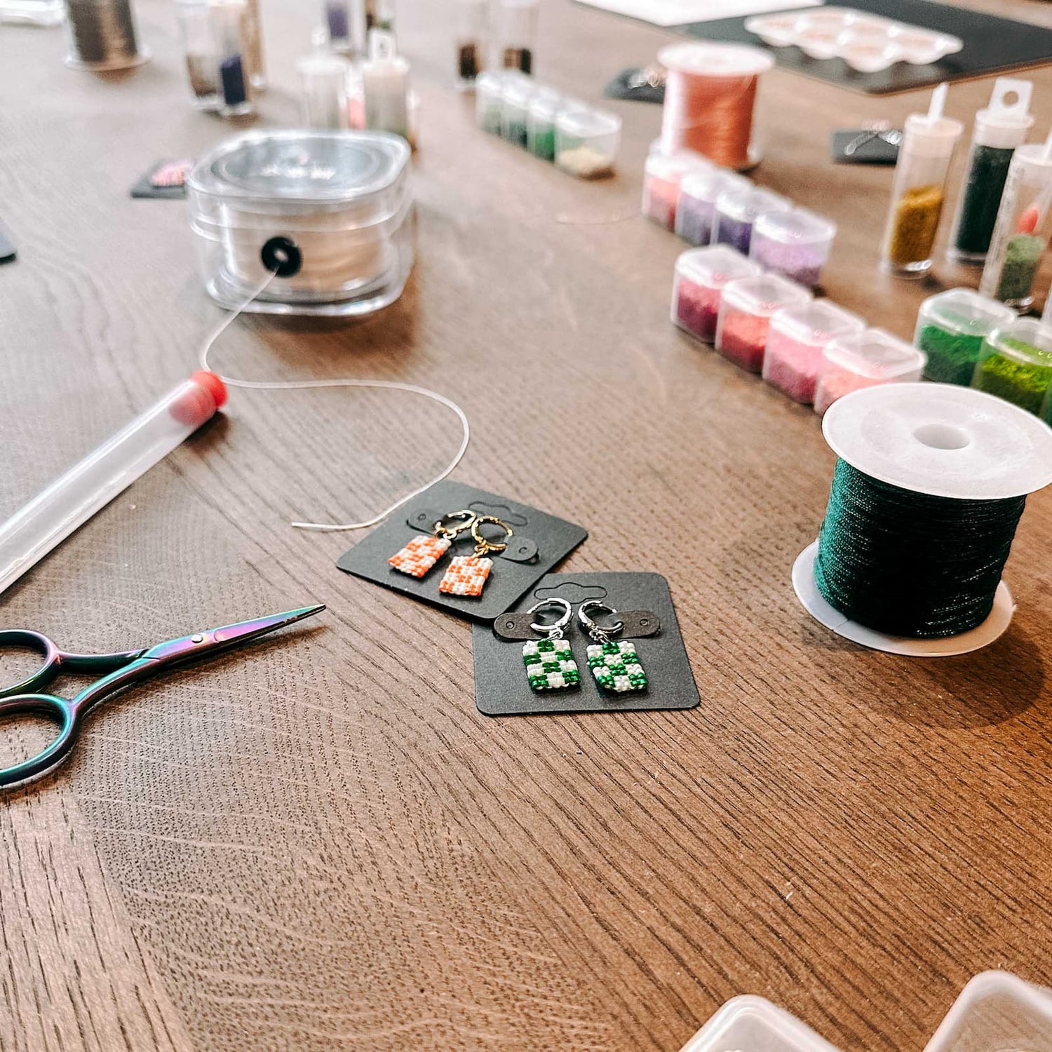 Perlenbänder im Hintergrund, vorne ein fertiges Paar Ohrringe und eine Schere auf dem Tisch beim DIY Perlenschmuck Workshop von GRIMMS GARTEN, Kreativkurs Berlin