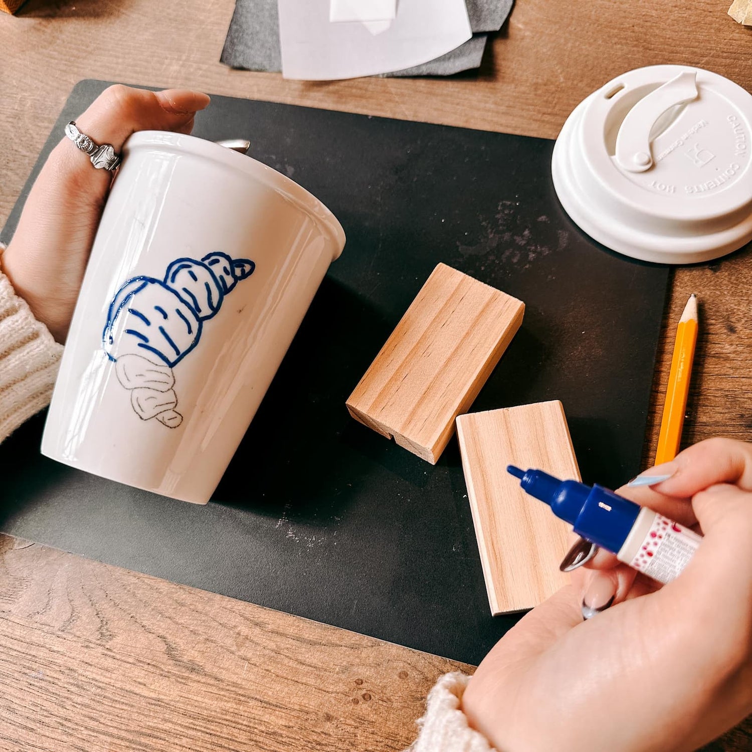 Hand zeichnet Croissant-Skizze mit blauem Stift auf Keramik To-Go Becher beim Keramik bemalen Workshop von GRIMMS GARTEN, Kreativkurs in Berlin
