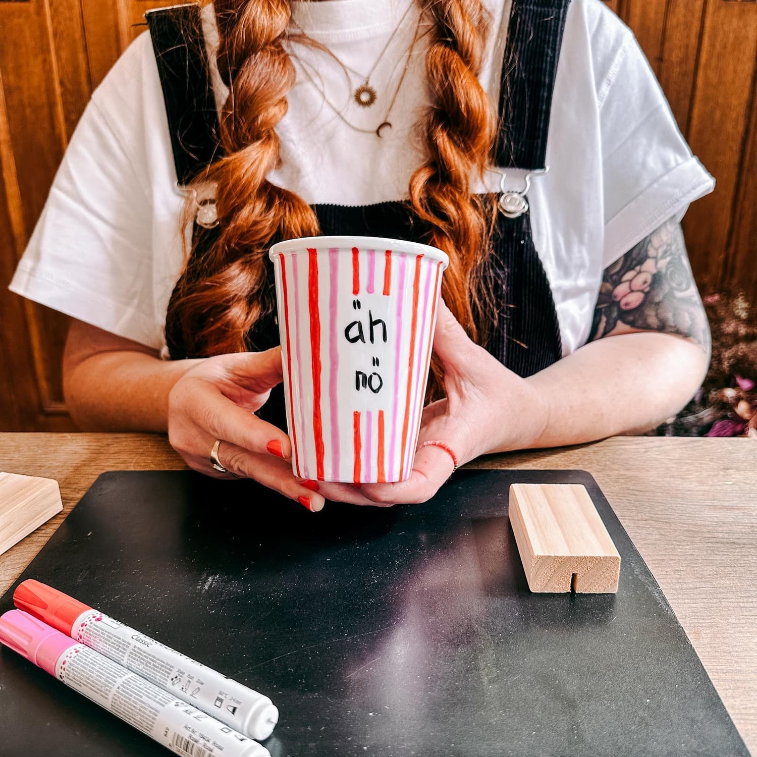 Hände halten Keramik To-Go Becher mit roten und rosa Streifen und Schriftzug beim Keramik bemalen Workshop von GRIMMS GARTEN, Kreativkurs in Berlin