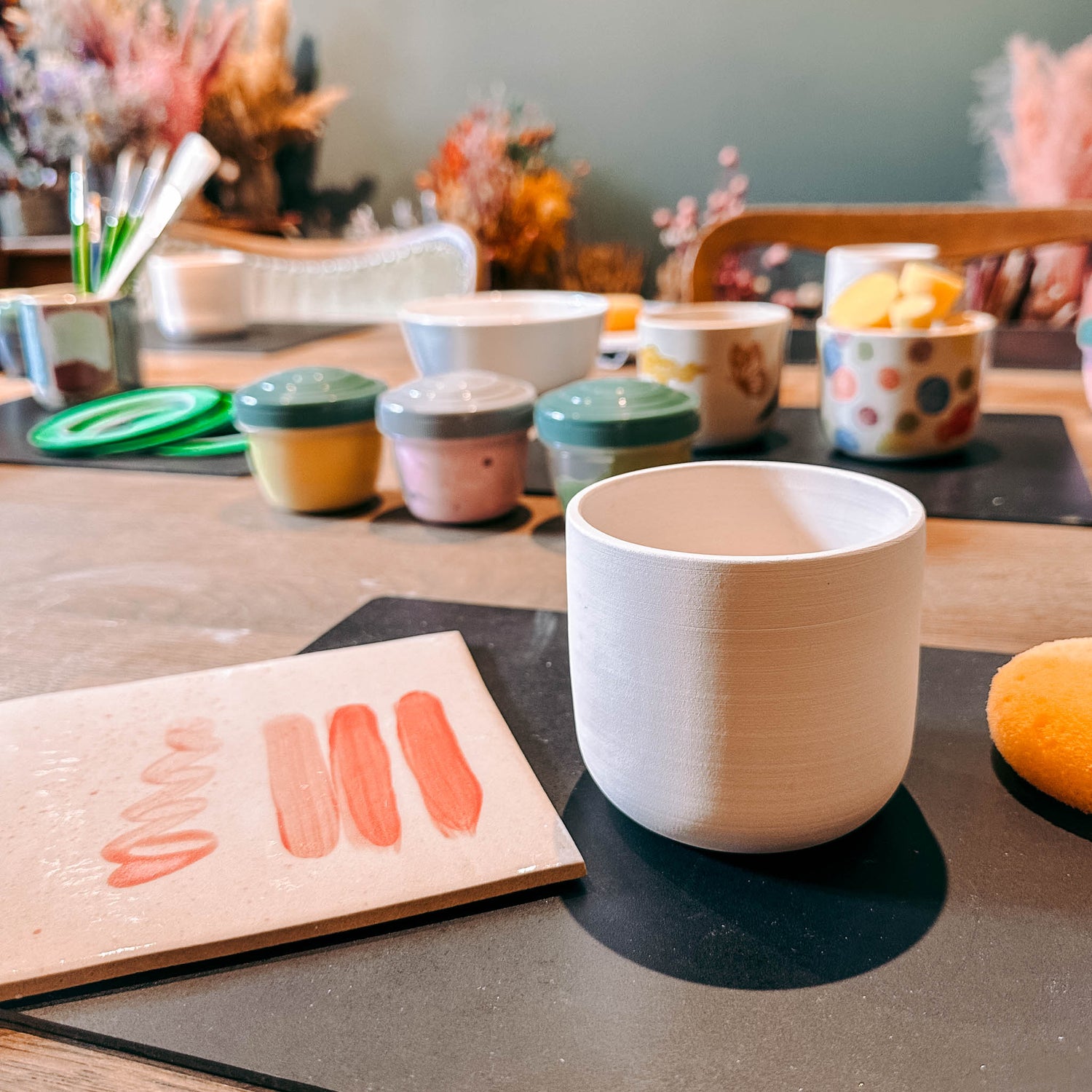 Weißer Keramikbecher auf Tisch mit Brett und pinken Farbswatches – DIY Keramik Workshop Berlin, kreativ bemalen