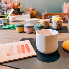 Weißer Keramikbecher auf Tisch mit Brett und pinken Farbswatches – DIY Keramik Workshop Berlin, kreativ bemalen
