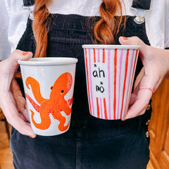 Hände halten zwei Keramik To-Go Becher mit bunten Motiven, einer mit Streifen und Schriftzug, der andere mit orangefarbener Krake beim Keramik bemalen Workshop von GRIMMS GARTEN in Berlin