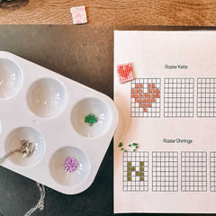 Grüne und lila Schmuckperlen in einer Schale neben Papier-Vorlagen mit Buchstabe N und Herz beim DIY Perlenschmuck Workshop von GRIMMS GARTEN in Berlin