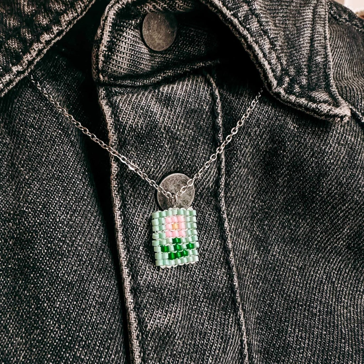 Nahaufnahme einer Perlenkette mit rosa Blumenanhänger vor grünem Hintergrund, getragen auf grauem Jeanshemd aus dem DIY Perlenschmuck Workshop von GRIMMS GARTEN in Berlin