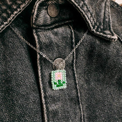 Nahaufnahme einer Perlenkette mit rosa Blumenanhänger vor grünem Hintergrund, getragen auf grauem Jeanshemd aus dem DIY Perlenschmuck Workshop von GRIMMS GARTEN in Berlin