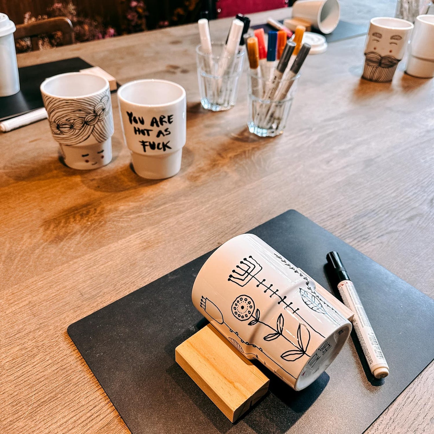 Keramik To-Go Becher mit Blumenwiesen-Motiv, schwarzer Stift daneben und weitere bemalte Becher im Hintergrund beim Keramik Workshop von GRIMMS GARTEN in Berlin